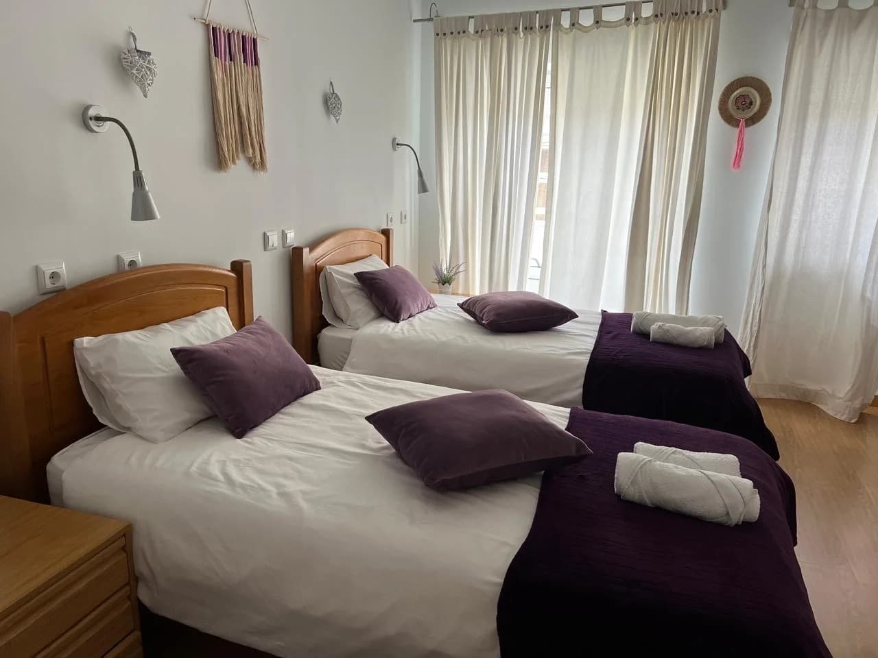 Quarto luminoso com duas camas individuais, roupa de cama branca, almofadas e mantas em tons ametista e cortinas claras com luz natural.