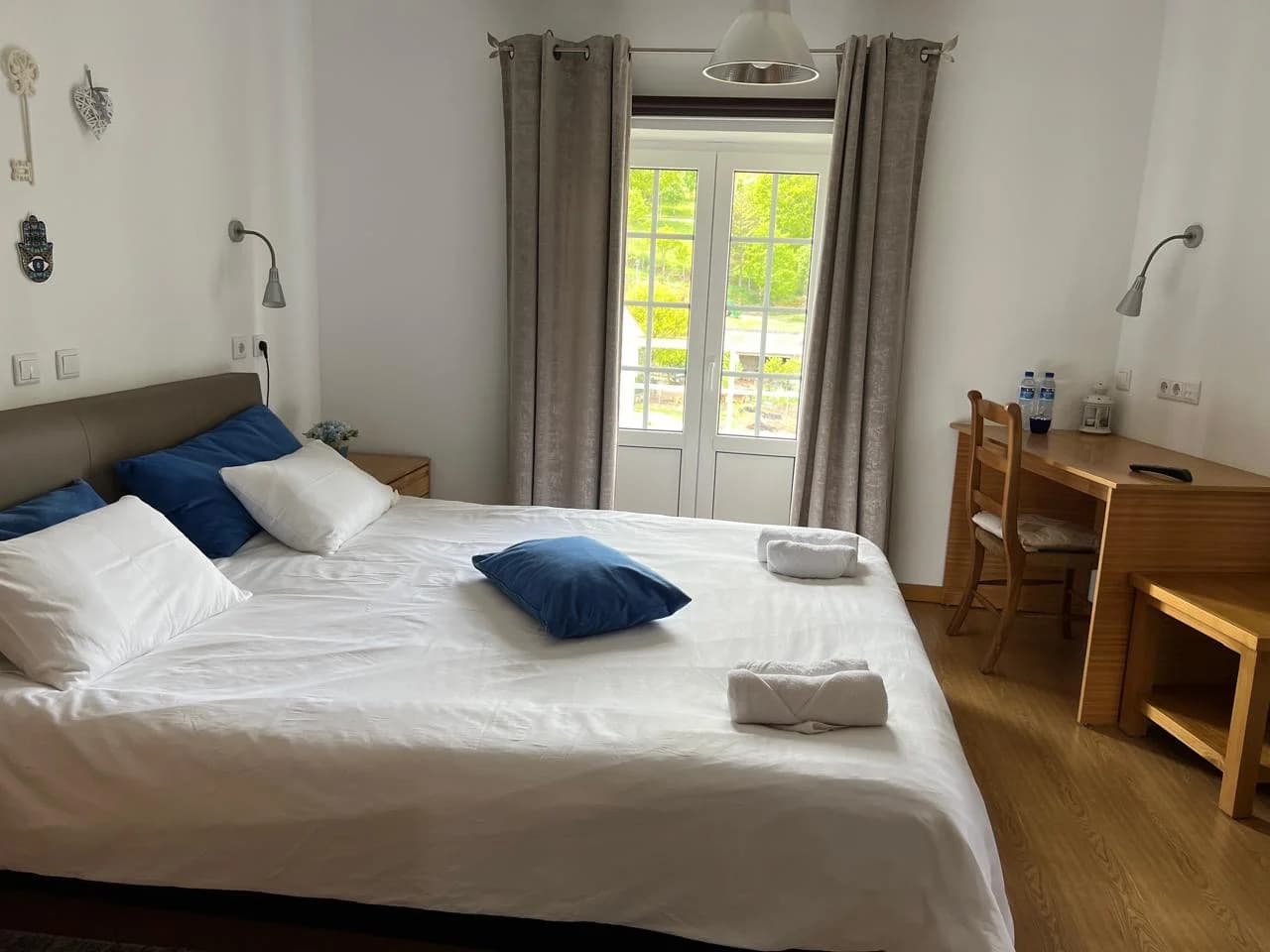 Quarto luminoso com cama de casal, roupa de cama branca, almofadas azuis, mesa de madeira e varanda com luz natural.